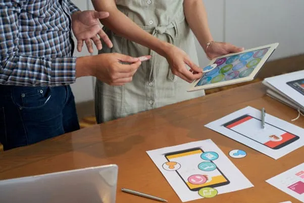 Team reviewing printed mobile app UI mockups alongside a tablet on a desk